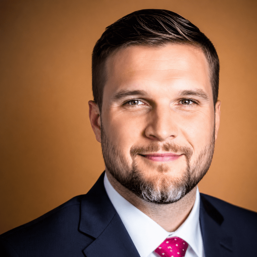 An image of a man in a navy suit with a red tie. He has brown hair and a short beard.